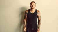 man with short hair and tattoos on his arms stands against a plain background. He is wearing a black tank top and looking directly at the camera with a neutral expression.